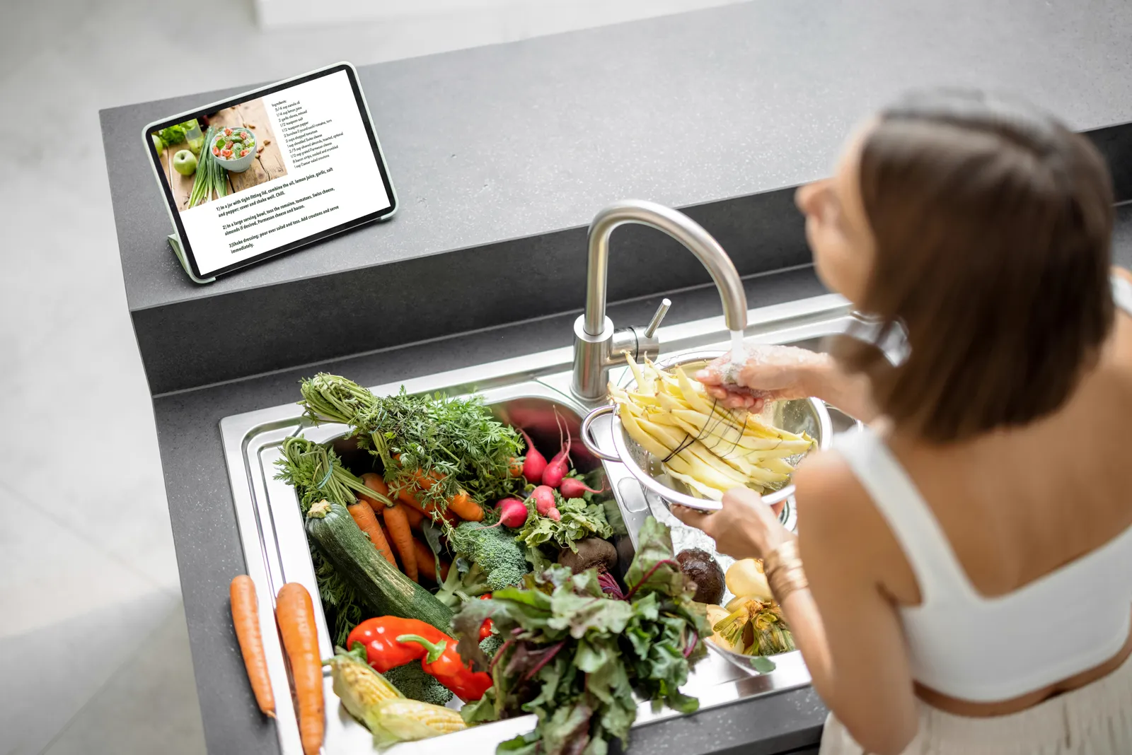 Overhead view of a woman washing fresh vegetables — carrots, radishes, potatoes — in a kitchen sink while a recipe sits open on a tablet nearby, illustrating how much of our daily minerals come from cooked food, not just drinking water